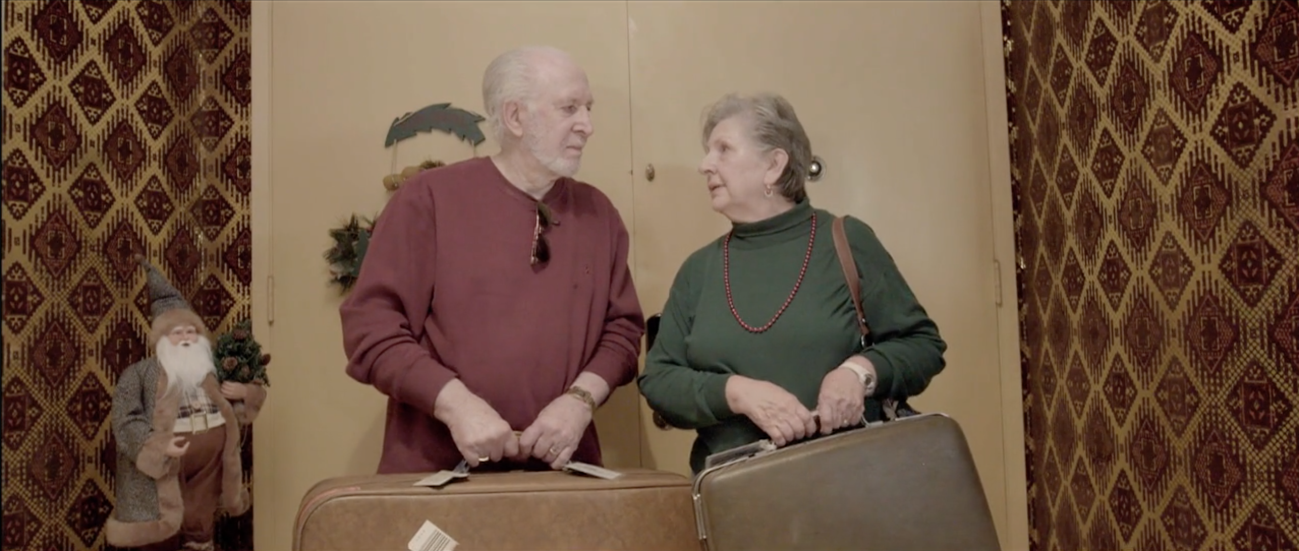 Older couple with suitcases before red patterned wallpaper, Takanakuy