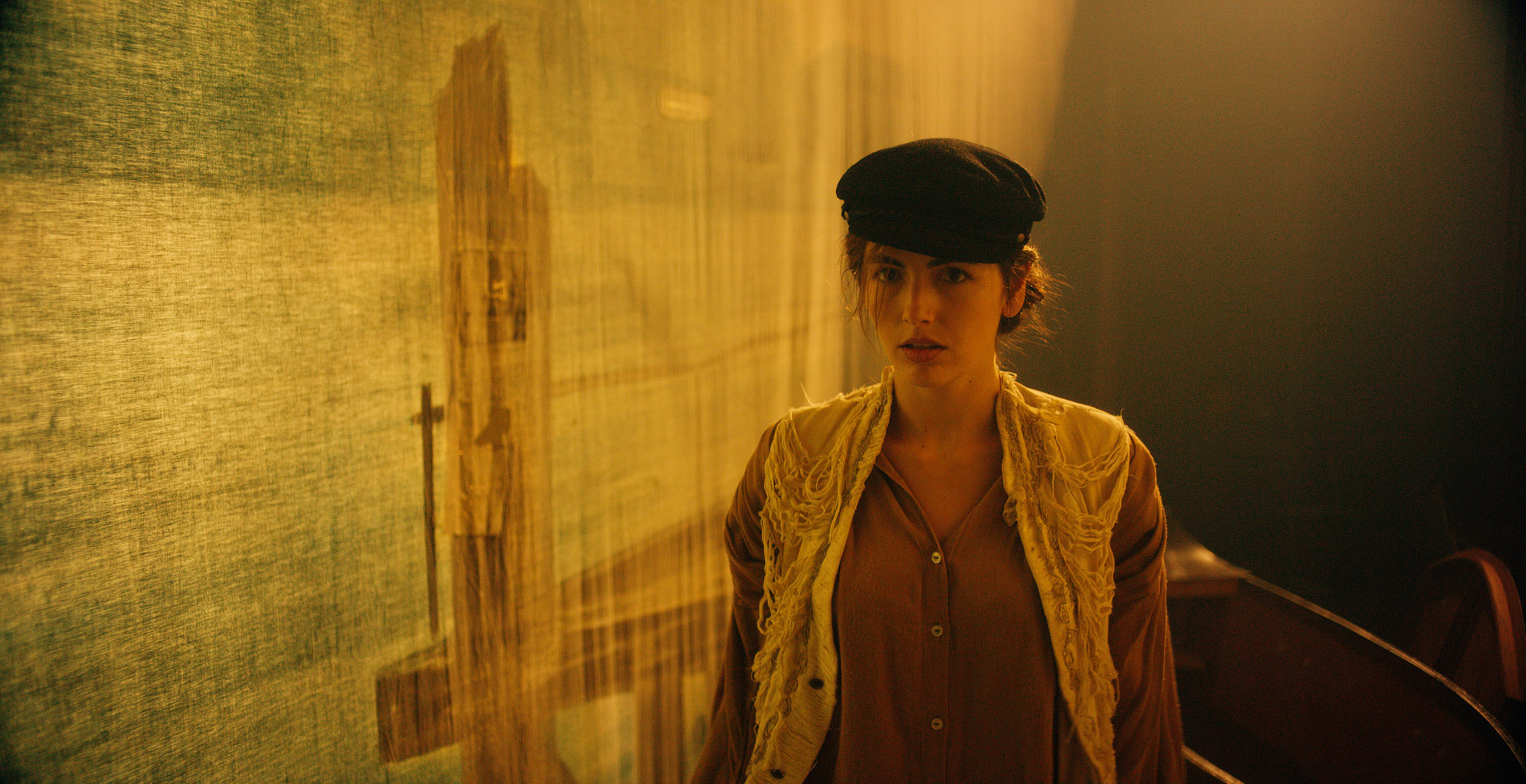 Character in a flat cap and olive jacket in a sunlit room, period dressing from The Mad Whale