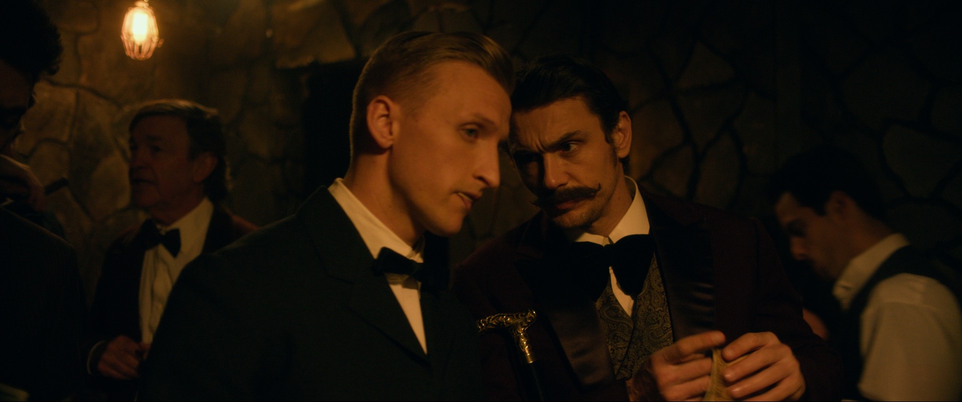 Men in Victorian formal dress in close conversation against a stone wall, The Mad Whale